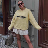 Woman wearing a yellow sweater with 'EME STUDIOS' text, standing on a street.